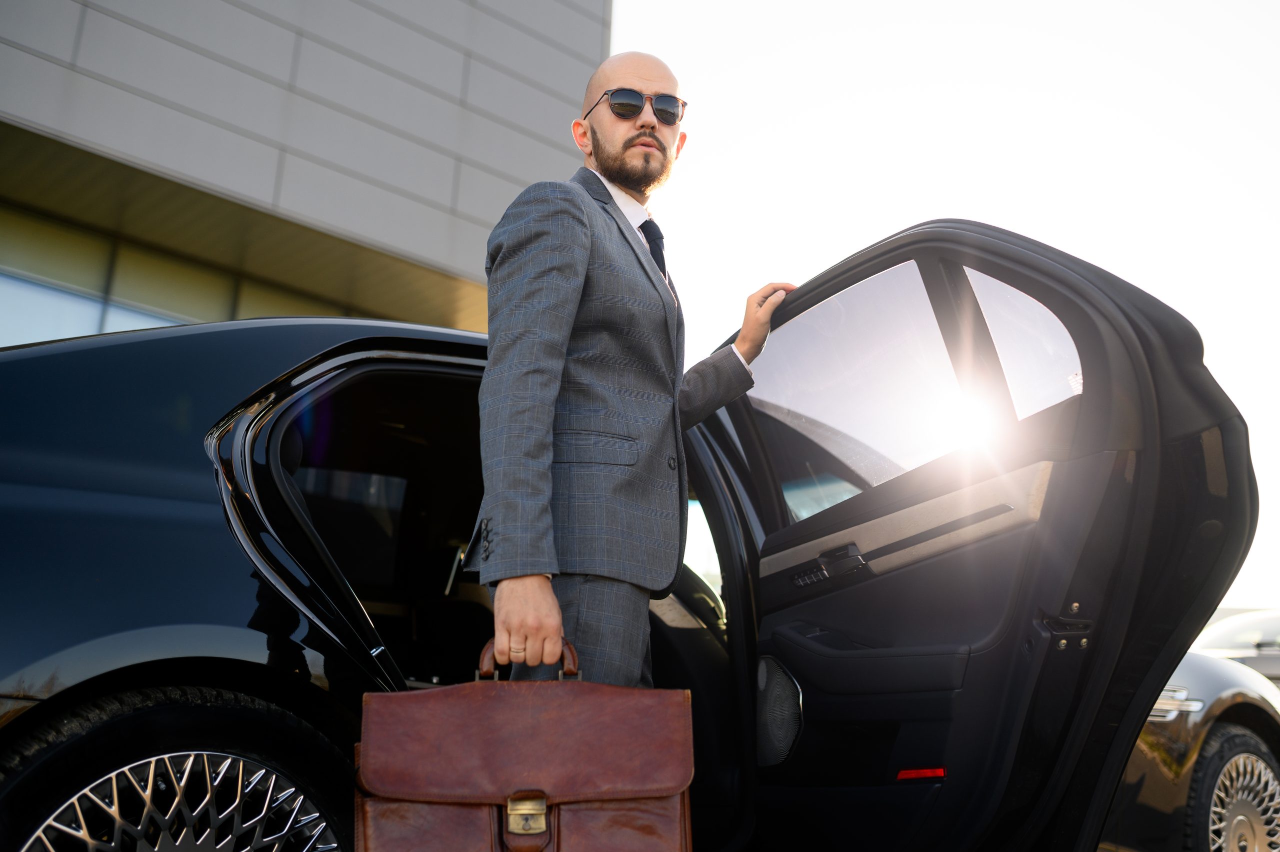 Portrait of a handsome businessman in sunglasses standing near the car outdoors in front of the modern building facade.